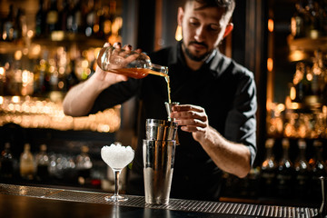 Male bartender pouring an orange alcoholic drink from the bottle to a steel jigger