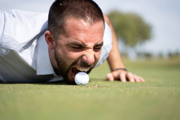 Man lying on a golf course biting a golf ball next to a hole