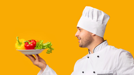 Cook Guy Holding Plate With Vegetables Over Yellow Background, Side-View