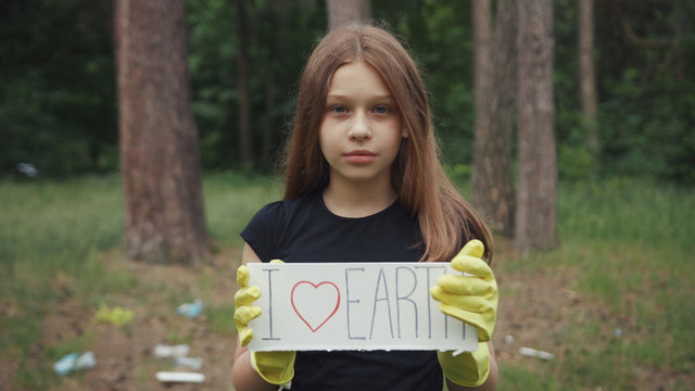 Outdoor portrait of a pretty serious girl protesting agsint earth pollution. Cute schoolkid holding a love earth sign staying in the summer nature park.