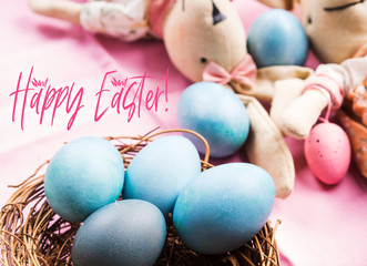 Pink easter holiday concept with bunny and blue colored hard boiled eggs in a wicker nest. Greeting card Happy Easter text