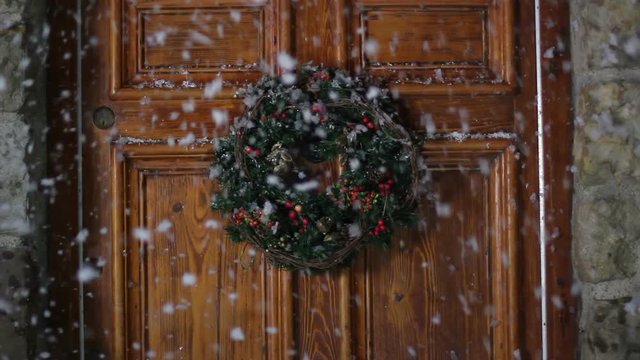 Christmas wreath hung on a front door and it's snowing. The snow falls outside the house at night.