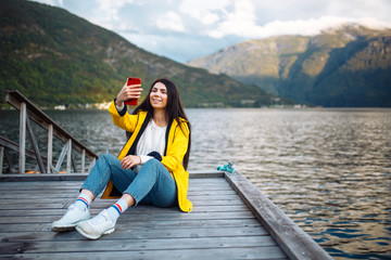 Fototapeta na wymiar The girl tourist takes a photo on the phone by the lake in Norway. Young woman takes selfie against the backdrop of the mountains in the Norway. Travelling, lifestyle, adventure, concept.