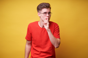 Keep secret! Handsome young man showing silence sign with his finger on lips. Studio portrait with yellow background.