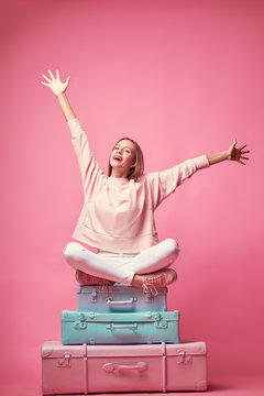 Ready for vacation. Traveling concept. Young excited woman sitting on the luggage valises. Pink background.