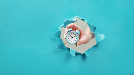 Hand holds alarm clock through a paper hole. Time management and deadline concept - obrazy, fototapety, plakaty