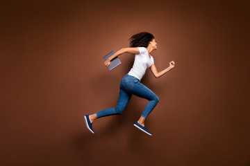 Fototapeta na wymiar Full length profile photo of funny dark skin lady jumping high holding notebook hurry enter classroom wear casual white t-shirt jeans isolated brown color background