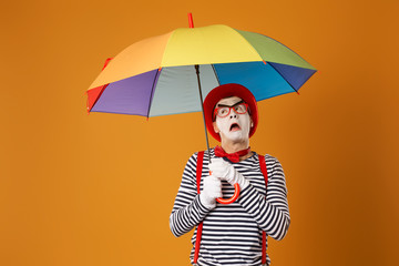 Surprised mime with multi-colored umbrella on empty orange background