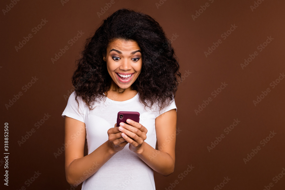 Sticker Portrait of amazed crazy afro american girl blogger use cellphone get social network notification look scream wow omg wear white t-shirt isolated over brown color background