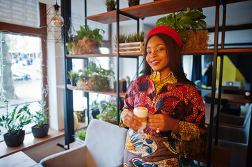Enthusiastic african american woman in trendy coloured outfit with red beret chilling in cozy cafe with cup of cappucino at hands.