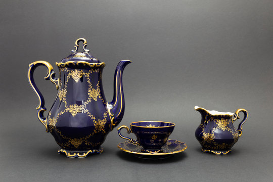 Closeup of a beautiful cobalt blue colored vintage porcelain tea set with golden floral pattern on dark gray background. The set includes a tea pot, a milk jug and a tea cup.