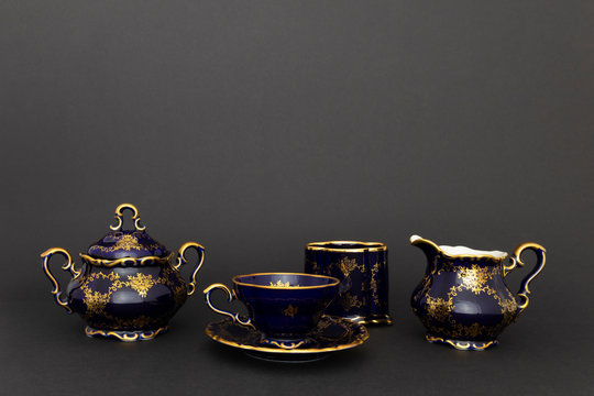Closeup of a beautiful cobalt blue colored vintage porcelain tea set with golden floral pattern on dark gray background. The set includes a a sugar bowl, a milk jug and a tea cup.