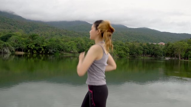 Slow motion of a young fit Asian woman running during her morning exercise at a local park with lake and mountain view, healthy lifestyle concept