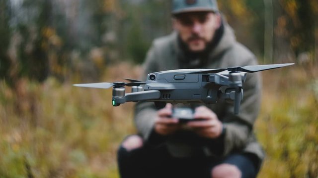 Close-up drone camera aircraft is controlled by young happy casual bearded Caucasian videographer man in autumn forest.