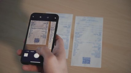 Hands of adult man holding modern smartphone and taking photo of bill invoice in special application. Confident male sitting behind wooden table inside workspace or bright light office - Powered by Adobe