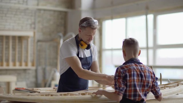 Middle aged father and little son in aprons building small wooden boat together standing at workbench in woodworking shop