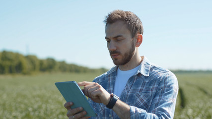 Serious concentrated young man using and typing on tablet outdoors. Farmer monitoring harvest in the green wheat field in summer.