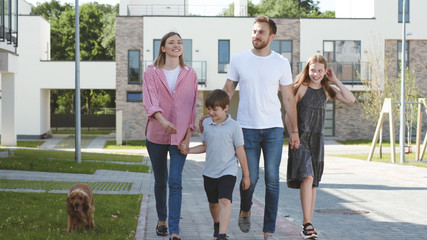 Happy affectionate young family walking in the neighbourhood of their new house. Smiling parents and their children holding hands exploring the new place on a sunny day.