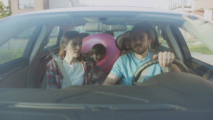 Front view of pretty married cuple and their kids riding a car in the city. Cheerful dad and his children giving high five having fun in the car during a family roadtrip to the seaside.