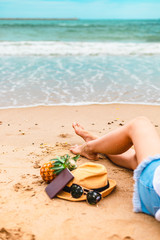 Woman tanned legs on sand beach. Sun glasses, pineapple, hat and passport on the beach and sea water flow.