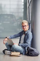 Young hipster guy sitting indoors near grey background with laptop and books