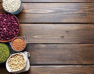 Bowls and pouches of various legumes: chickpeas, lentils, mung, red and white bean on a brown wooden background.