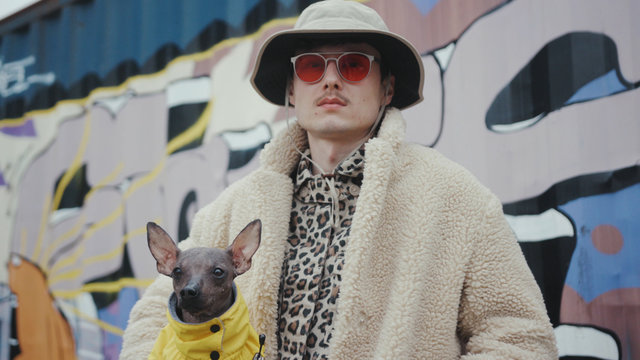 Portrait young man with hat in red sunglasses holding dog wear yellow clother in arms look at camera serious fashion style background artwall fun pet small young adult expression slow motion