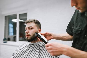 Professional barber makes a beard haircut for a client with a clipper. Hairdresser makes beard correction to handsome man. Man does a haircut at a men's hairdresser and looks into the camera.