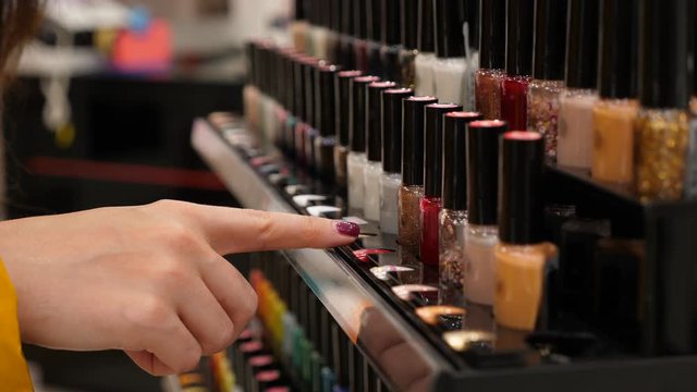 Woman choose nail polish at beauty store, close up shot. She put pointer finger against varnish samples, watch and and think what to take, then pick up red one
