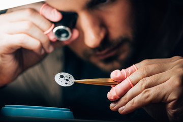 Selective focus of watchmaker holding eyeglass loupe and watch part in tweezers