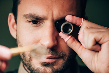 Selective focus of handsome watchmaker holding eyeglass loupe and watch part
