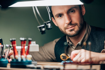 Handsome watchmaker looking at camera while working with watch parts and magnifying glass