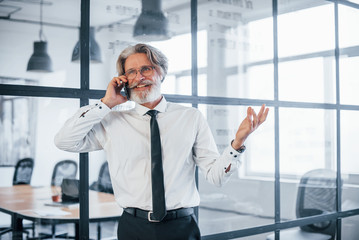 Mature businessman with grey hair and beard in formal clothes have conversation by the phone in the office
