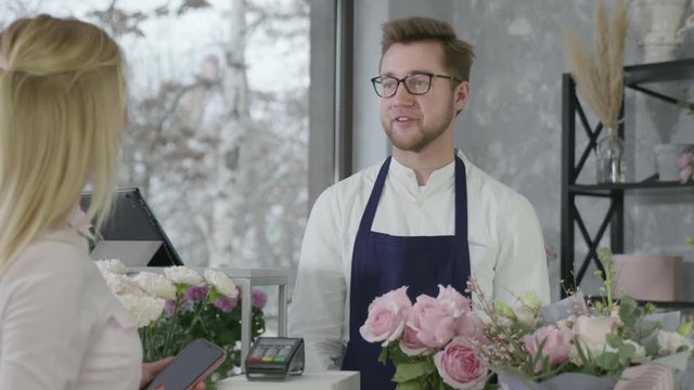 young businessman, male startup small business owner of modern flower shop sells beautiful bouquet of flowers to gerl, woman pays for purchase by contactless payment using smartphone