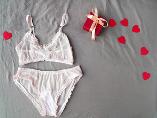 Red gift box, hearts and women's pink lingerie at grey bed as background