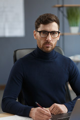 Young businessman working in office with laptop. Business portrait of handsome bearded man wearing eyeglasses sitting at workplace. Confident businessman became successful. Business concept.