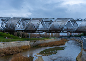 Obraz premium Views of the Arganzuela Bridge on a cloudy day, pedestrian bridge crossing the Manzanares River in the Madrid Río Park, formed by a metal spiral covered with a mesh, travel concept