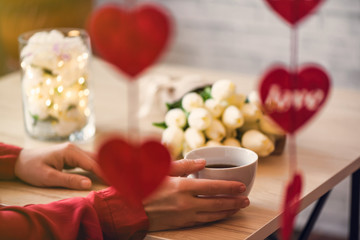 Valentines day concept. Cropped of lonely woman sitting on the table drinking coffee with bouquet of flowers tulips celebrating in restaurant. View through a heart-shaped garland.