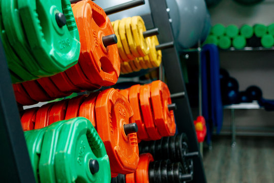 Dumbbells Of Different Sizes. Dumbbells In The Fitness Room. Dumbbells Of Orange, Green And Yellow. Rack With Metal Pancakes Of Different Weights.