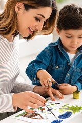 Fototapeta premium happy woman sitting at table and drawing with paints together with son