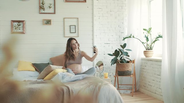 Beautiful expecting mother taking selfie photo. Smiling pregnant woman in bed.
