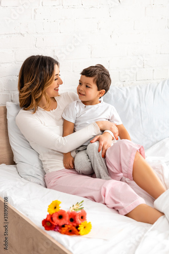 cheerful mom and son looking at each other while sitting in bed near flowers on mothers day