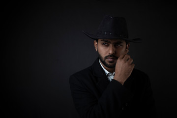 Fototapeta na wymiar Fashion portrait of an young and handsome Indian Bengali brunette man with striped formal shirt, black suit and cowboy hat in black copy space background. Indian lifestyle and fashion photography.