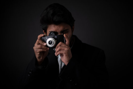 Fashion portrait of young Indian Bengali brunette man with striped formal shirt, black suit capturing photograph in camera in black copy space background. Indian lifestyle and fashion photography.