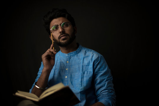 Fashion portrait of young Indian Bengali brunette man with traditional cotton wear and glasses holding pencil and book in black copy space background. Indian lifestyle and fashion photograph
