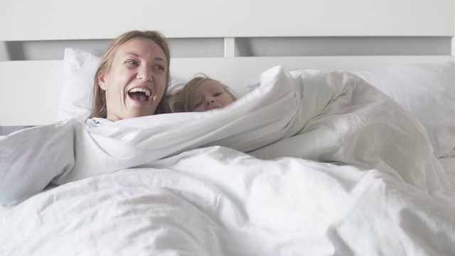 Mom and daughter hide under a blanket in the bed. Mom and baby peek out from under blankets and laugh cheerfully
