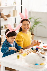 cheerful child smiling at camera while sitting at desk near white rabbit in wicker and painting Easter eggs together with cute brother