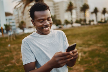 Portrait of a young male athlete standing at park smiling while texting messages on mobile phone