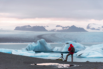 photographer wearing red jacket taken a photo at Jokulsarlon, Iceland with small iceberg floating in the lake and beautiful mountain and sky
