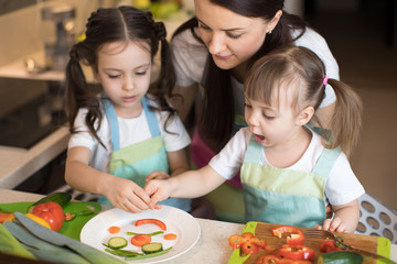 happy family mom and her little daughters are preparing healthy food, they make smiley face with vegetables morsels in domestic kitchen